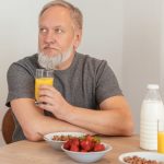 Elderly man with a beard enjoying a nutritious breakfast with orange juice, cereal, and strawberries.