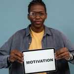 A young African American man holding a tablet displaying the word 'MOTIVATION' against a blue background.